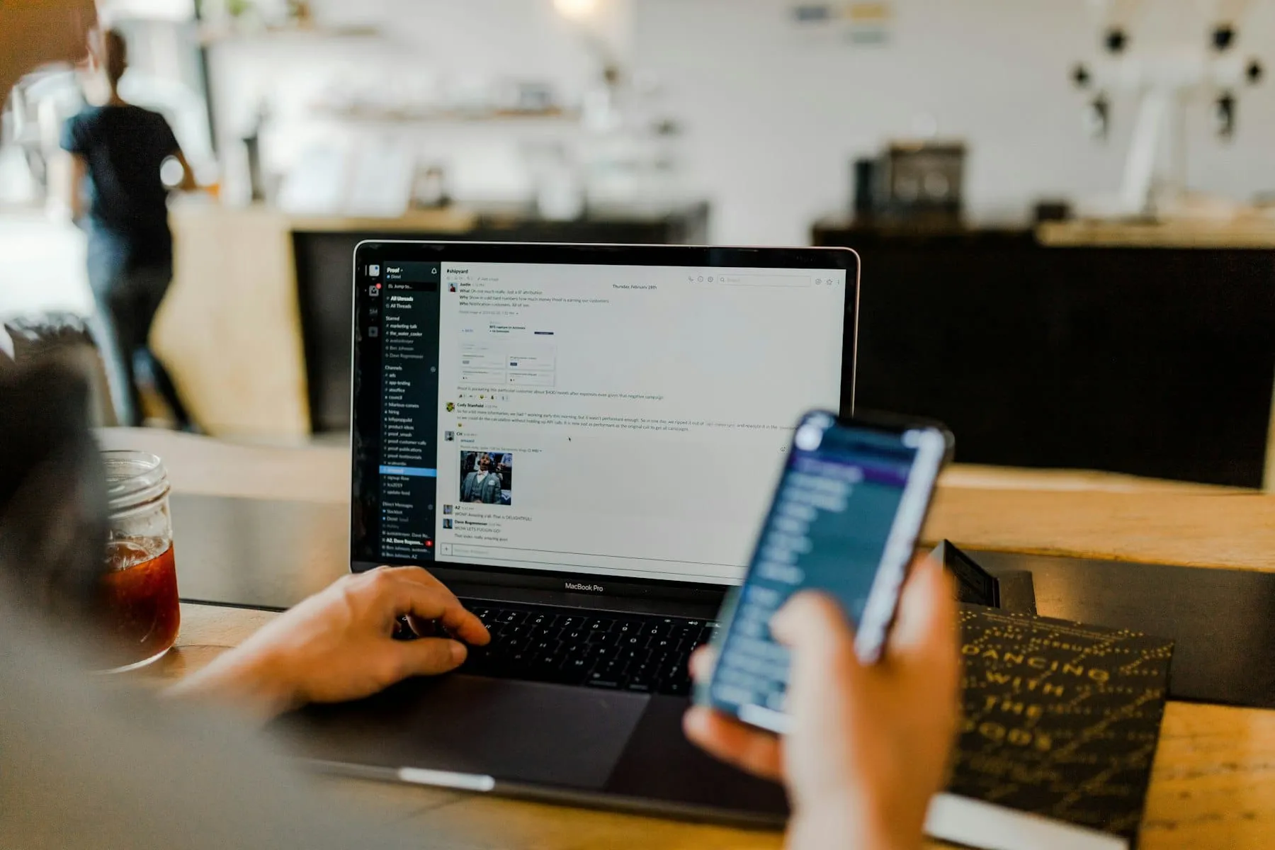 Iemand kijkt op een telefoon naast een laptop en een kop koffie