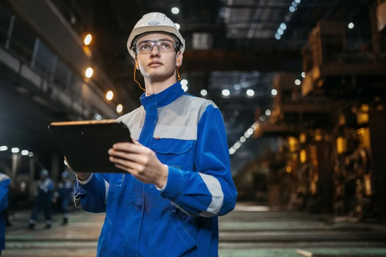 Een medewerker in blauwe werkkleding met helm en tablet in een fabriekshal